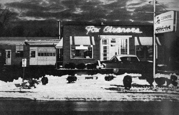 Black and white photo of "fox cleaners" store at dusk with vintage car parked in front, snow on ground.