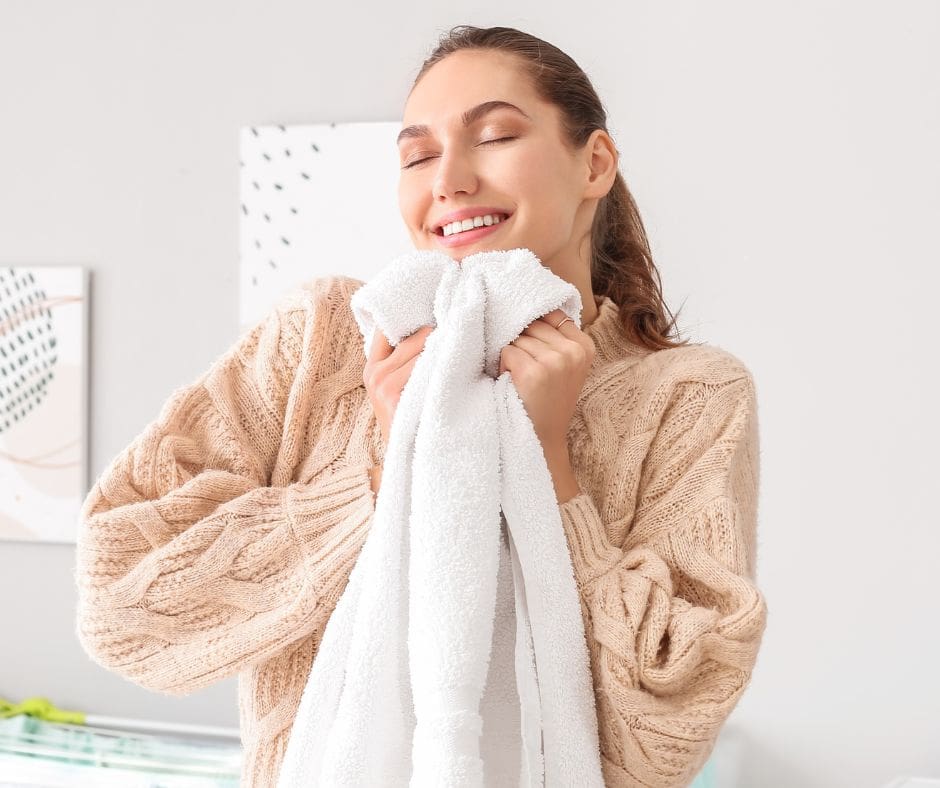 A woman in a beige sweater smiles with closed eyes, holding a white towel close to her face.