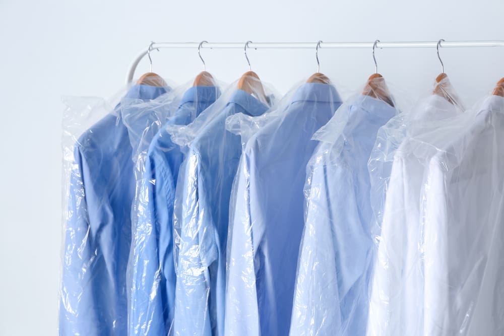 A row of shirts on hangers, each covered with clear or blue plastic garment bags, against a white background.