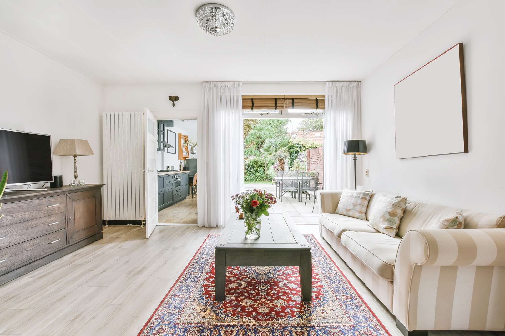 Bright living room with a striped sofa, wooden coffee table, red patterned rug, TV stand, and open view to the kitchen and garden patio through glass doors.