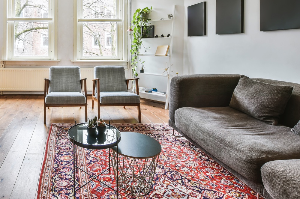 A modern living room with a gray sofa, two gray armchairs, round black coffee tables, a red patterned rug, wooden floor, and large windows with white walls and shelving.
