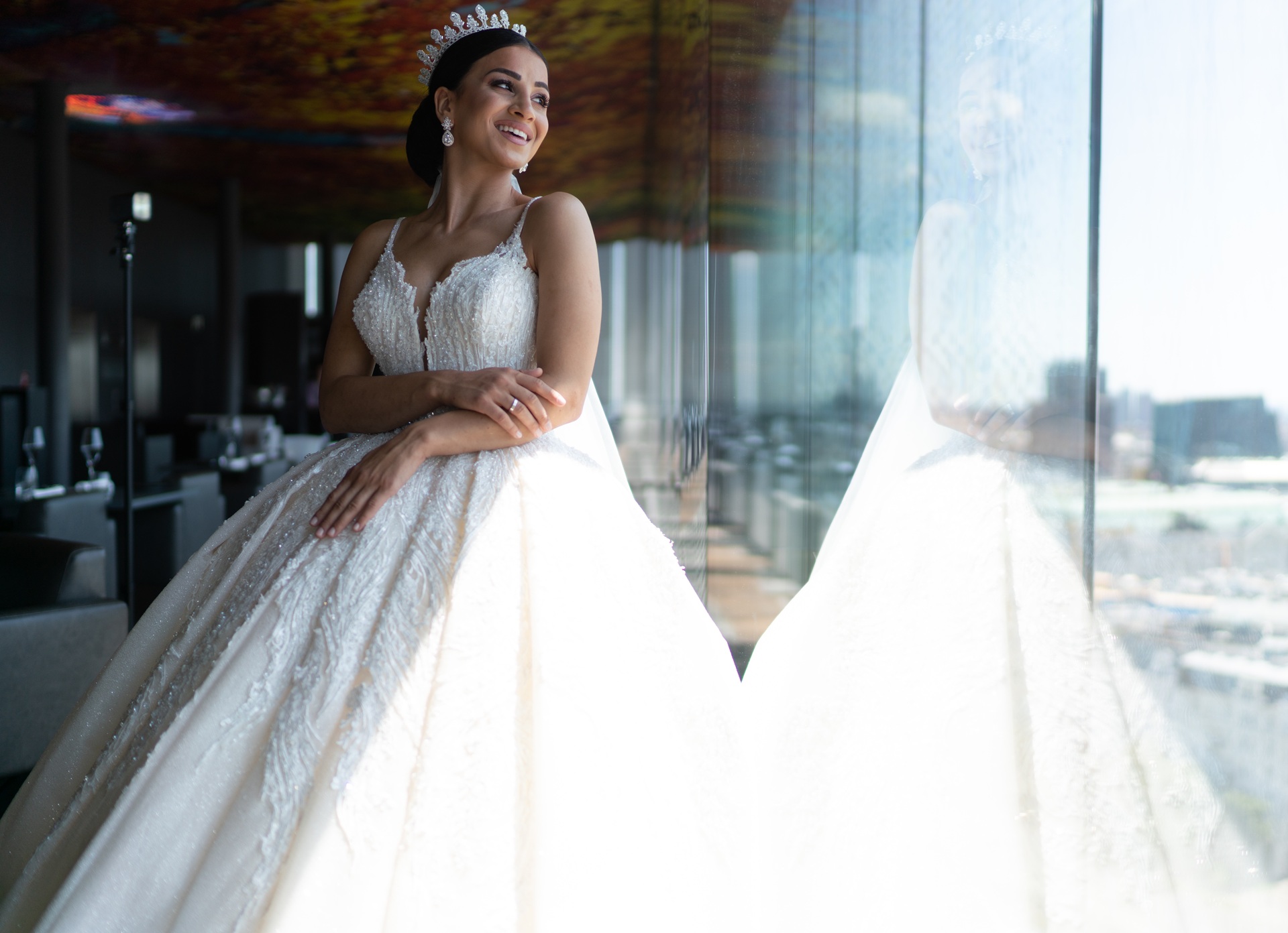 A woman in a white wedding gown and tiara stands by a large window, smiling, with her reflection visible in the glass.
