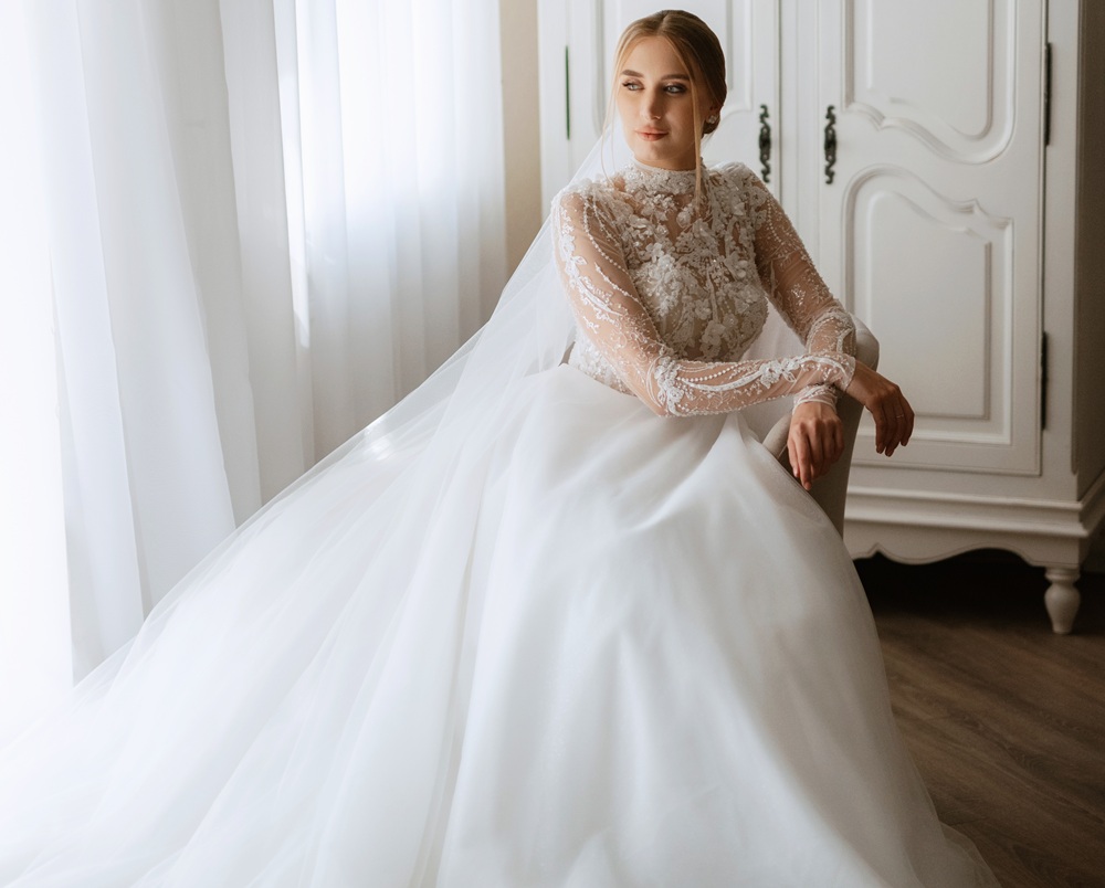 A bride in a long-sleeved lace wedding dress sits beside a window with sheer white curtains, looking to the side.