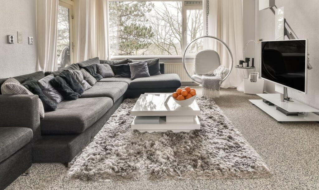 Modern living room with a gray sectional sofa, a shaggy rug, a white coffee table with a bowl of oranges, a hanging chair, and a wall-mounted TV near large windows.