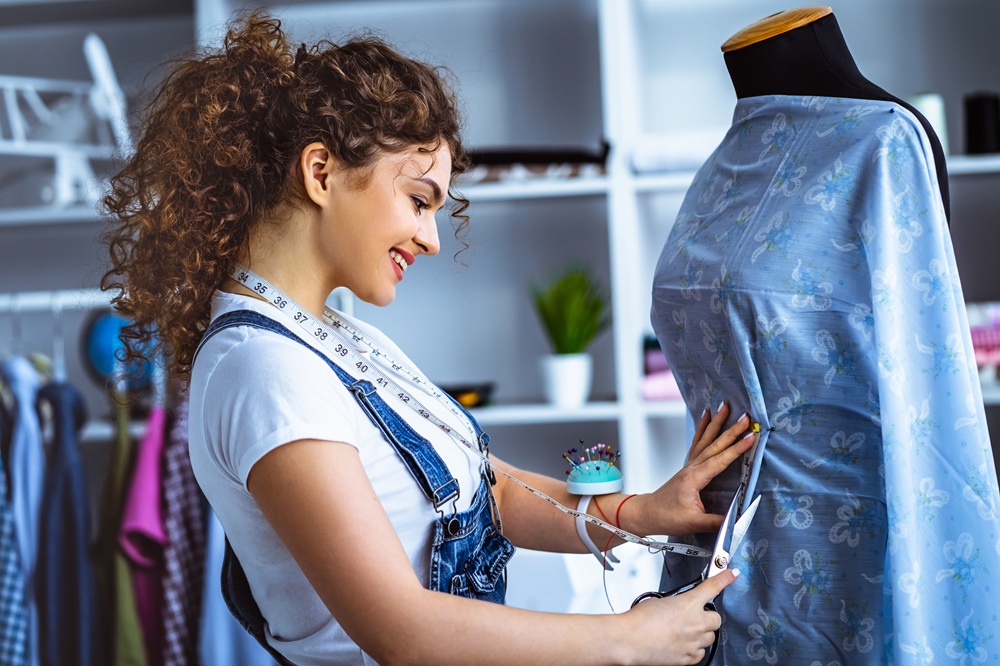 A woman with curly hair cuts blue patterned fabric on a dress form, wearing a white shirt, overalls, and a measuring tape around her neck in a workspace.