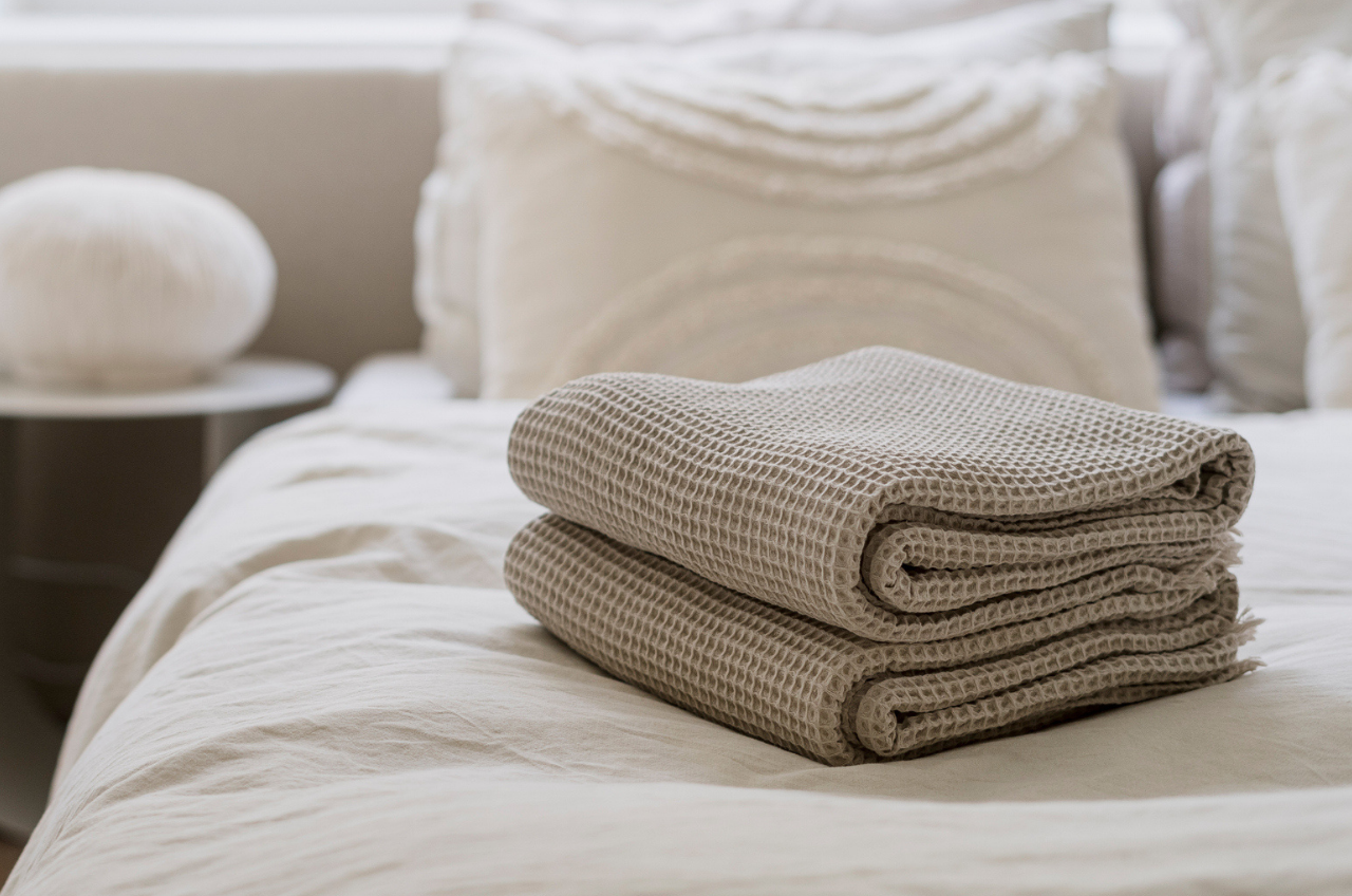 Two folded beige waffle-textured towels are placed on a neatly made bed with white linens and decorative pillows in the background.