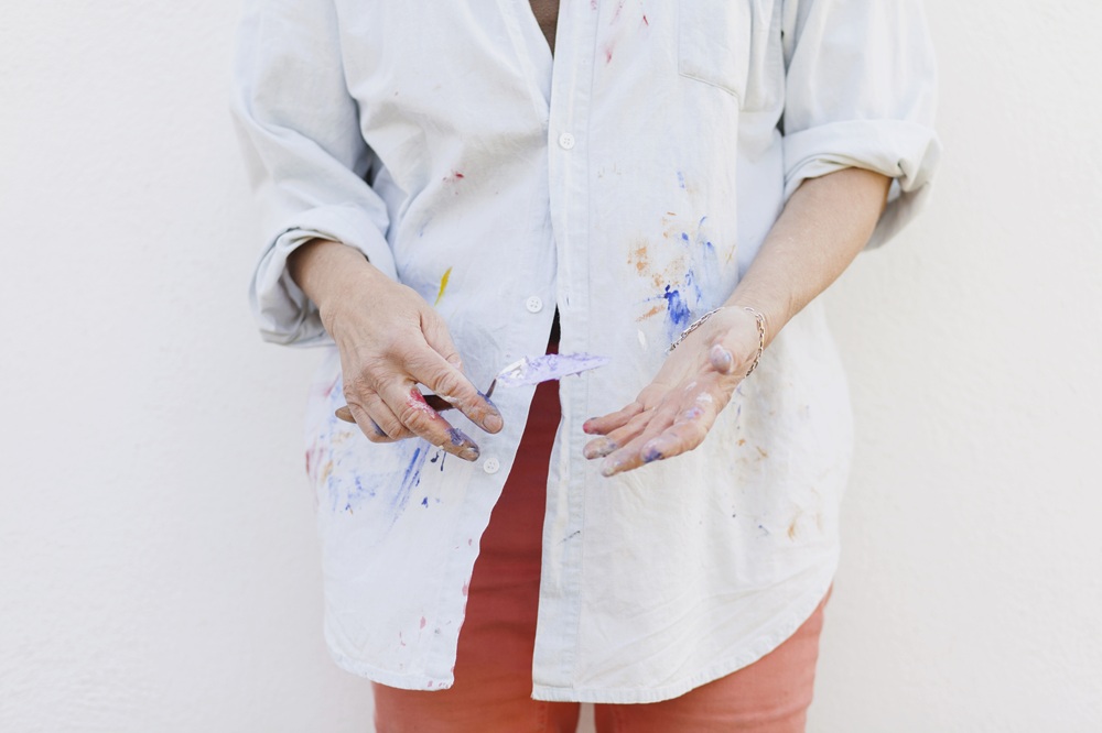 Person wearing a paint-stained button-up shirt and red pants holds a paintbrush in one hand and has paint on their other hand, standing against a plain white background.