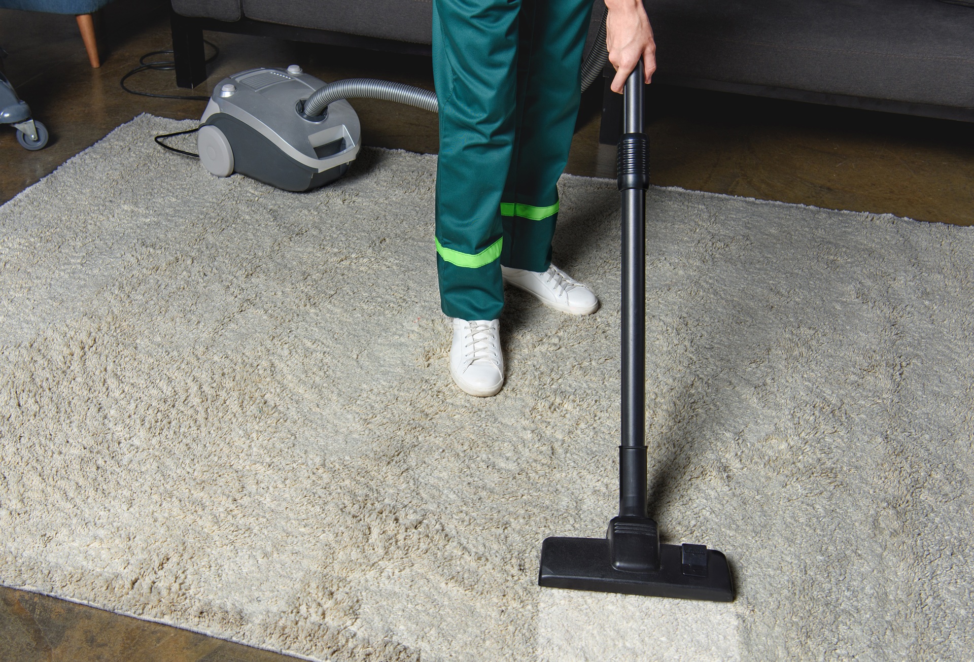 Person in green uniform vacuuming a light-colored rug with a canister vacuum cleaner, standing in front of a dark sofa.