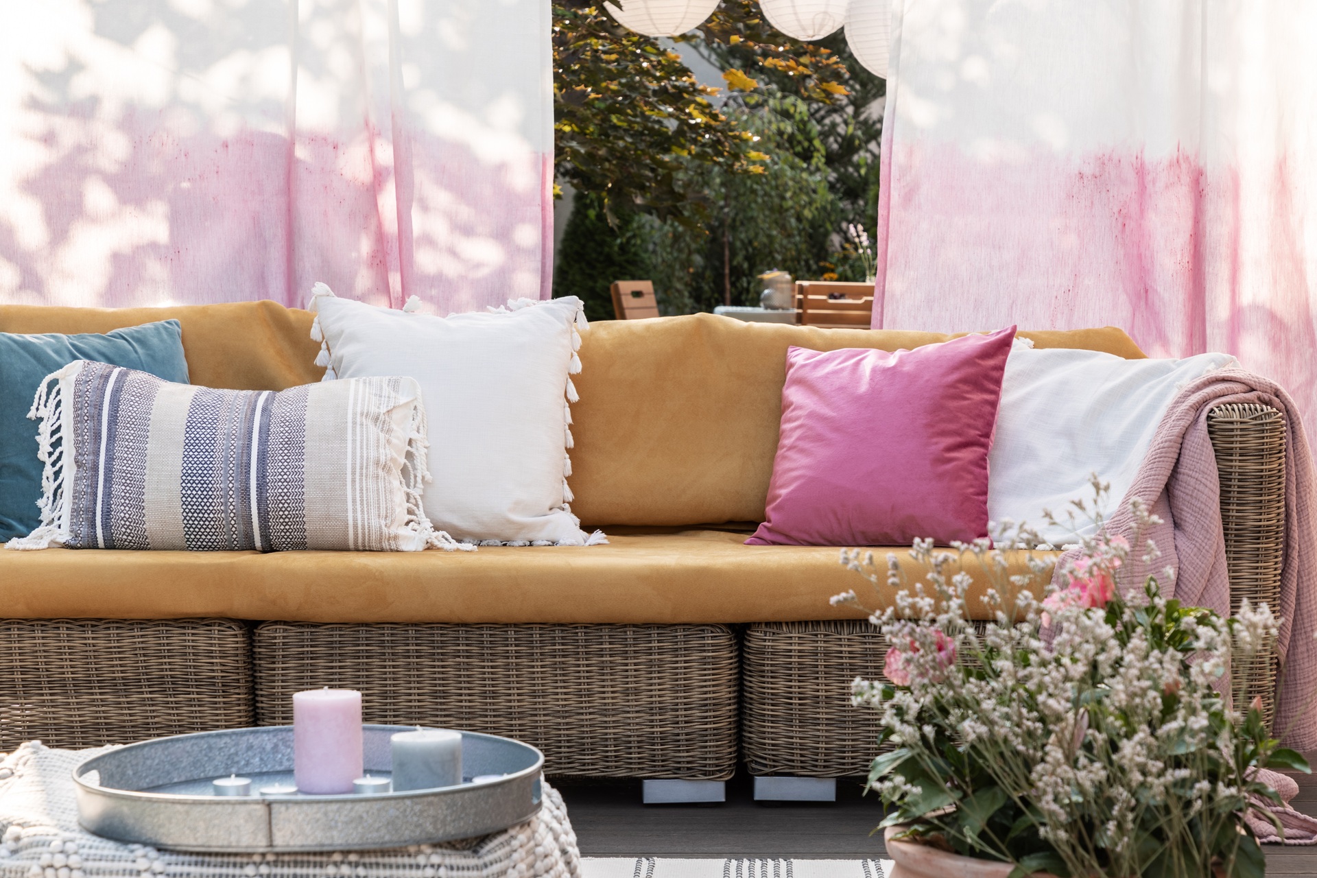 A wicker outdoor sofa with mustard yellow cushions and assorted throw pillows stands in front of pink-and-white curtains; a plant and candles sit on a nearby table.