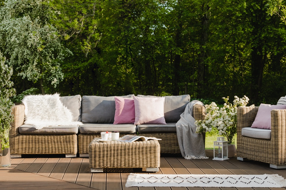 Wicker outdoor sofa and armchair with gray and pink cushions on a wooden deck, surrounded by greenery and decorated with flowers, a lantern, and a tray with cups.