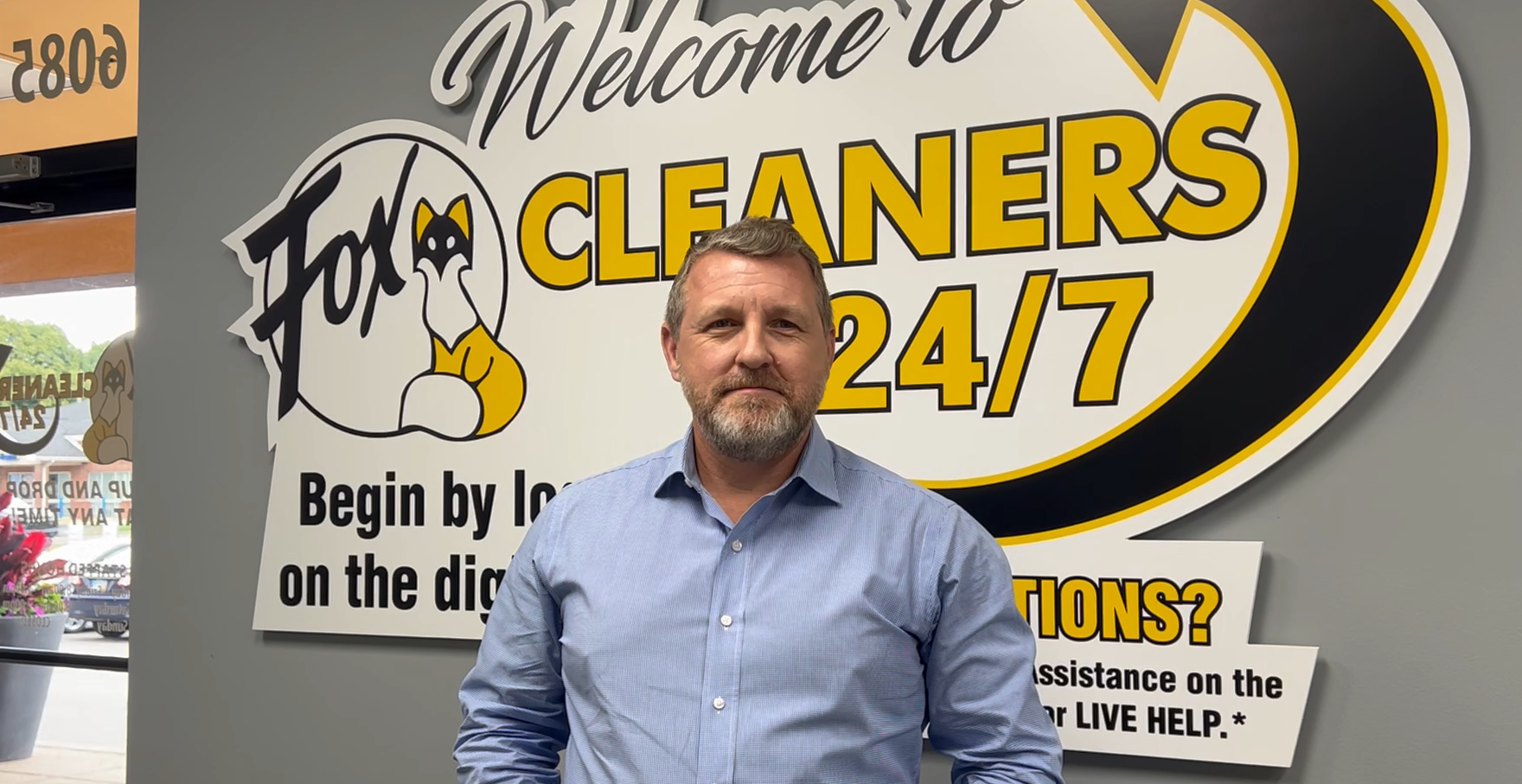 A man in a blue shirt stands in front of a sign that says "Fox Cleaners 24/7" inside the kiosk entrance.