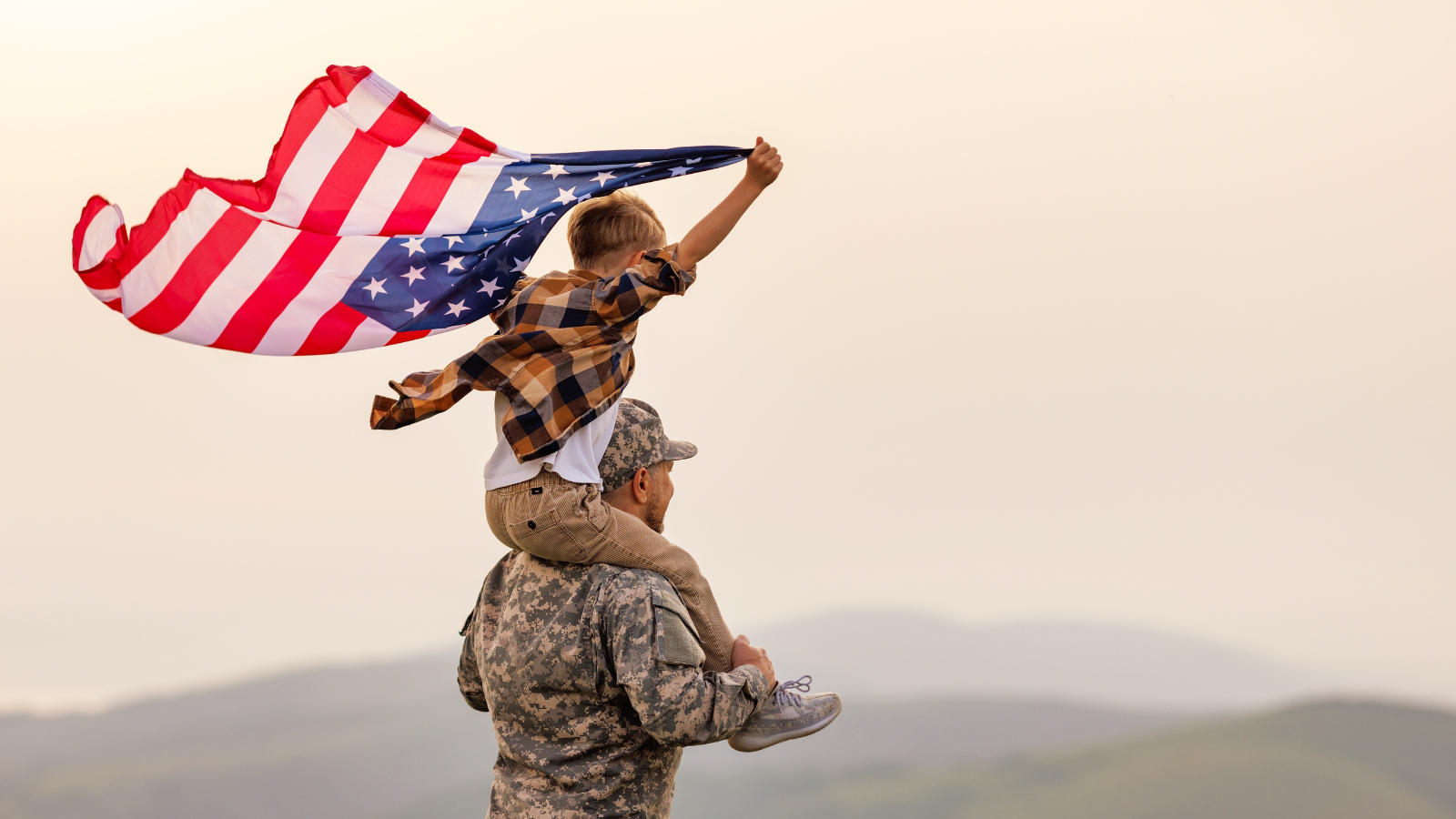 A child holds an American flag while sitting on the shoulders of an adult veteran in military uniform outdoors with hills in the background.