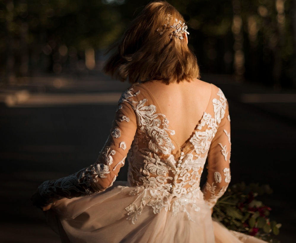 A woman with shoulder-length hair wearing a lace wedding dress with a low back stands outdoors, holding the skirt and a bouquet, with sunlight on her back.