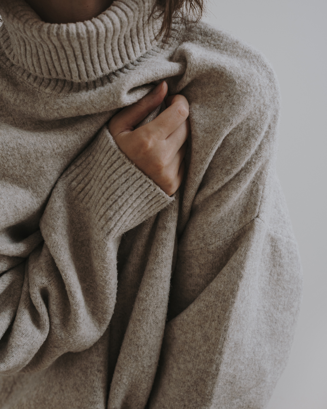 A person wearing a beige turtleneck sweater holds the fabric near their collarbone with one hand; their face is not visible. A person wearing a beige turtleneck sweater holds the fabric near their collarbone with one hand; their face is not visible.