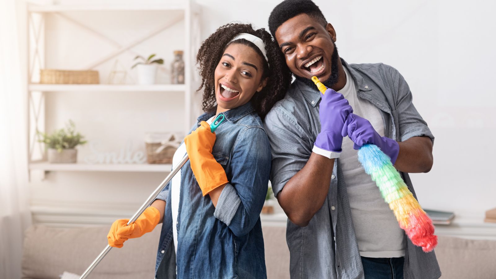Two people in casual clothes and rubber gloves smile and pose with cleaning tools, ready to spring clean a brightly lit living room.