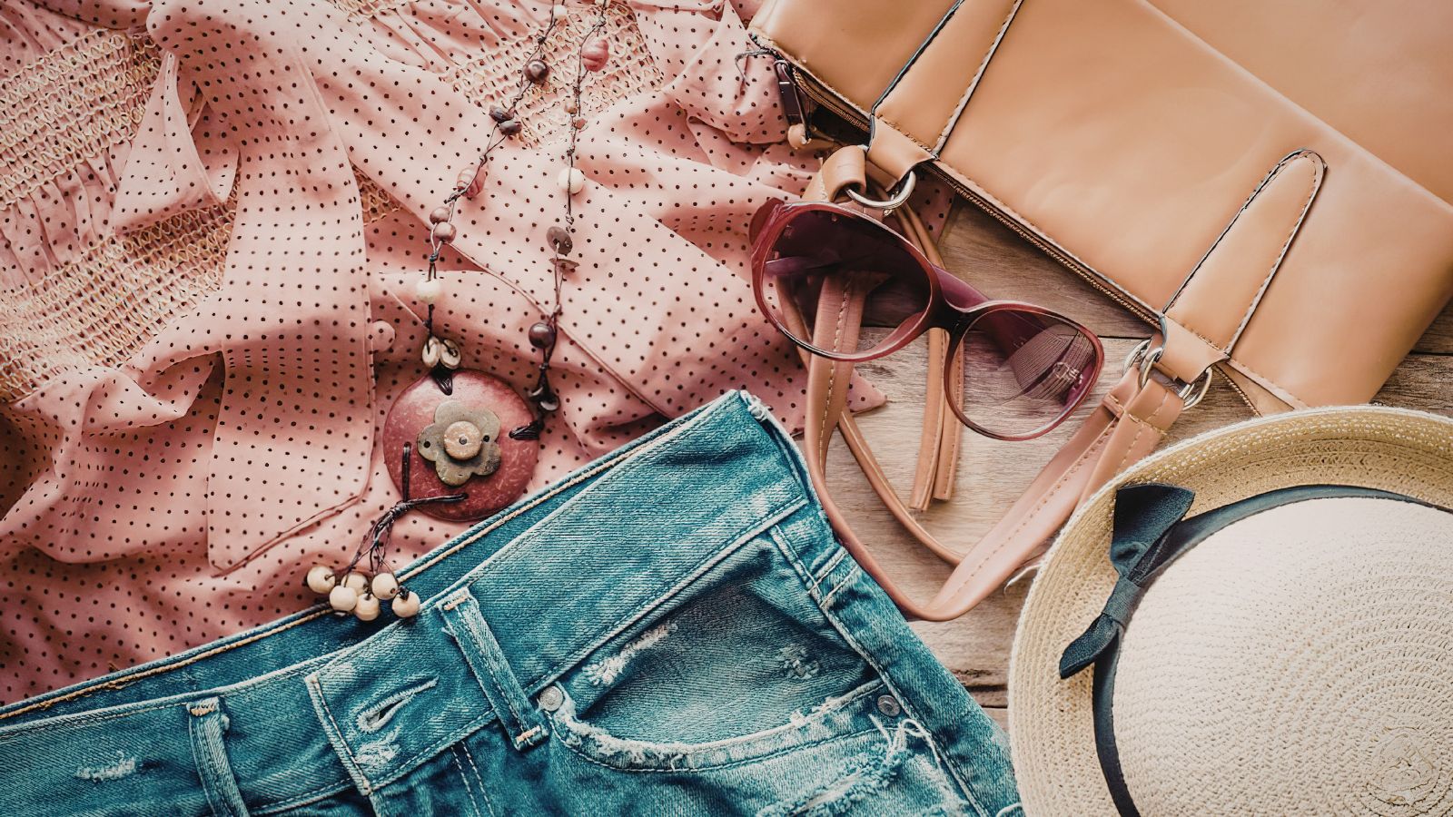 Summer style on display: a pink polka dot blouse, blue denim shorts, beaded necklace, brown sunglasses, tan handbag, and a straw hat with a black ribbon arranged on a wooden surface.