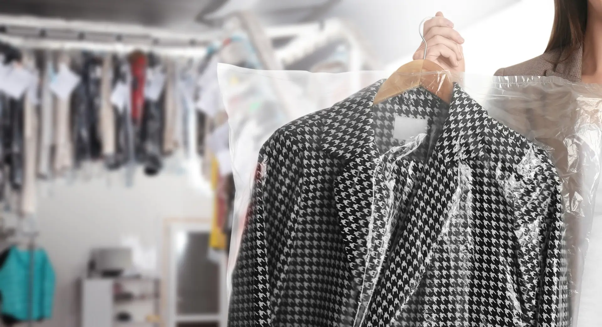 A person holds a houndstooth patterned coat on a wooden hanger, covered in plastic, with a clothing rack and garments visible in the background.