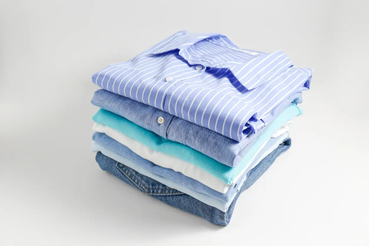 A neatly folded stack of blue and white shirts and jeans on a plain white surface. A neatly folded stack of blue and white shirts and jeans on a plain white surface.